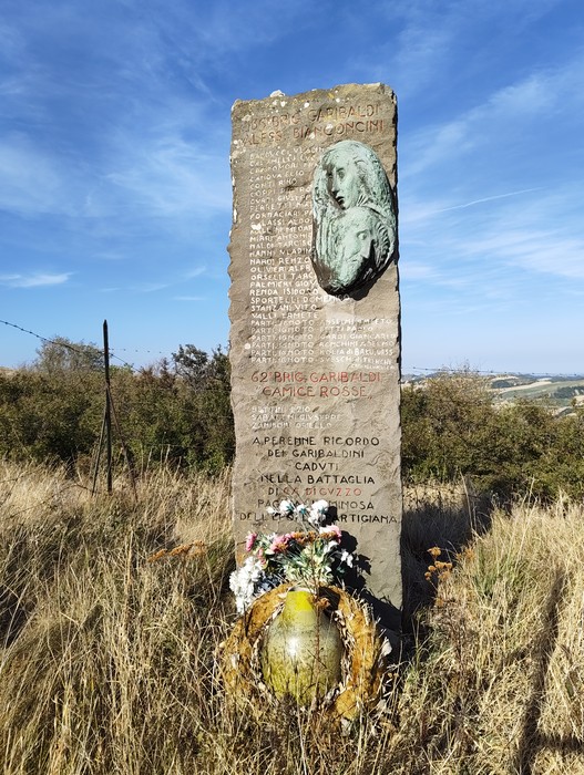 Stele in pietra con iscrizioni e rilievo di un volto, posta tra l’erba alta e la vegetazione collinare.