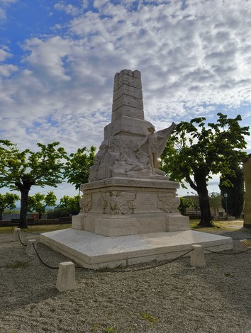 Monumento ai caduti di Piazze, con statue e un alto obelisco, immerso in un giardino verde e cielo nuvoloso.