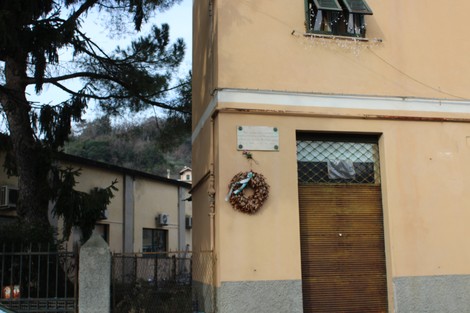 Lapide commemorativa su un edificio a Montecucco, con una corona di fiori appesa. Ambiente urbano circostante.