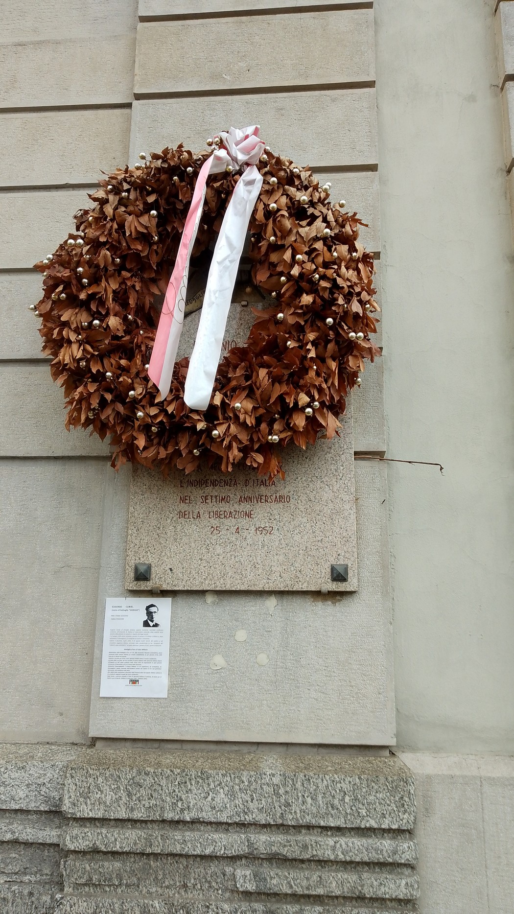 Lapide commemorativa con una corona di fiori secchi e nastri, apposta su una parete di pietra.