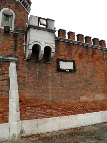 Lapide commemorativa su un muro di mattoni, adornata con una corona di fiori e decorazione tricolore.