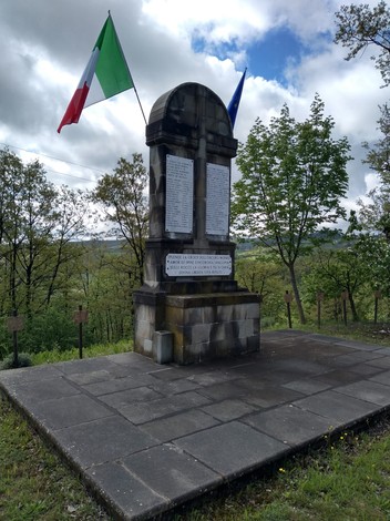 Monumento ai martiri dei Boschi di Ciano, circondato da alberi e bandiere italiane ed europee.