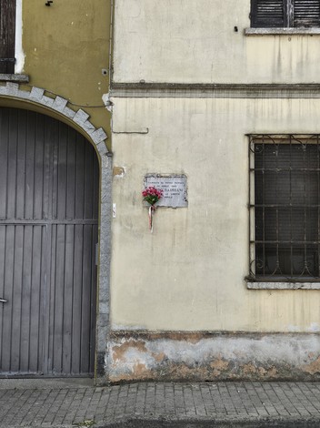 Lapide commemorativa affissa su una parete esterna, con fiori decorativi nei pressi.