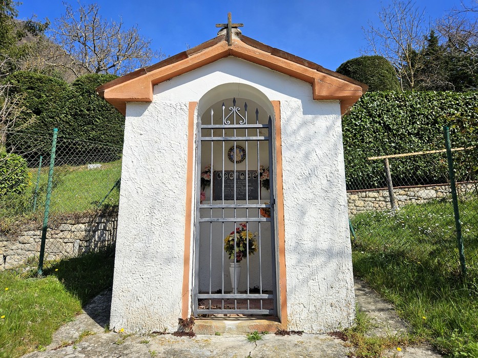 Cappella votiva bianca con tetto a due falde, croce sul frontone, cancello in ferro e corone all'interno.
