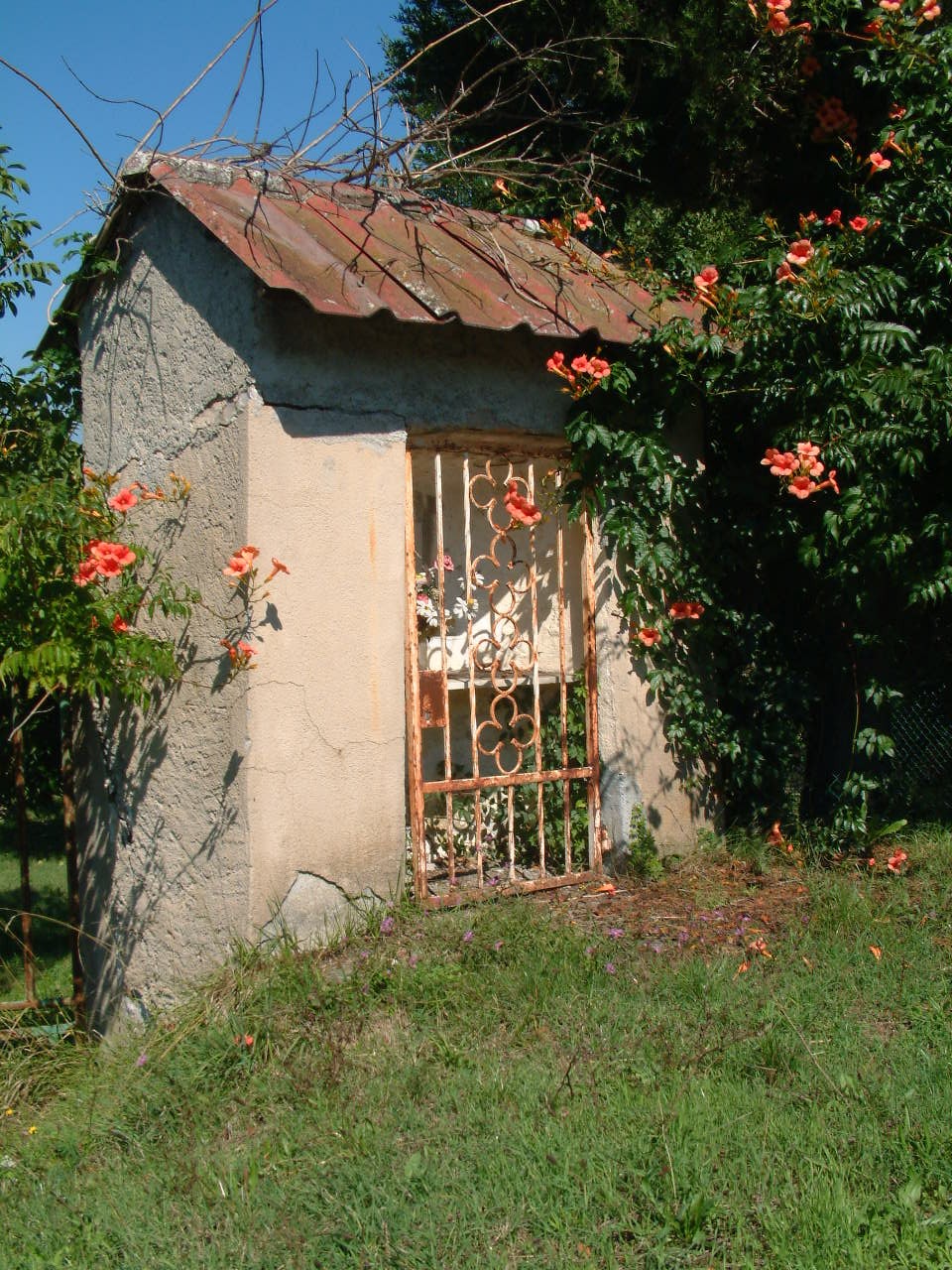 Tabernacolo commemorativo dell'eccidio di Canova, immerso nella vegetazione e circondato da fiori.