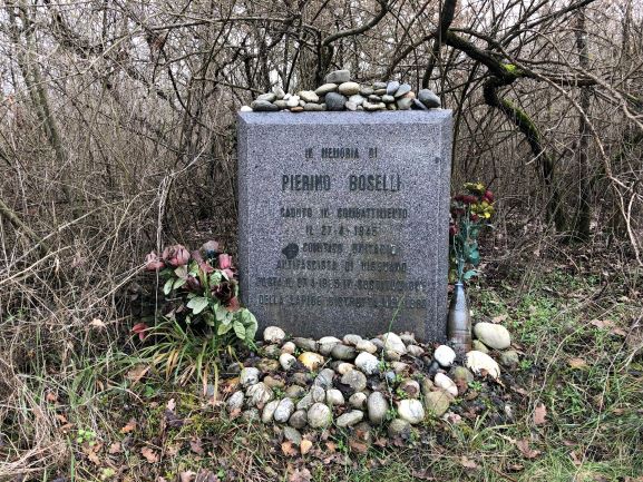 Cippo commemorativo di Pierino Boselli, adornato con fiori e sassi, immerso nella vegetazione.