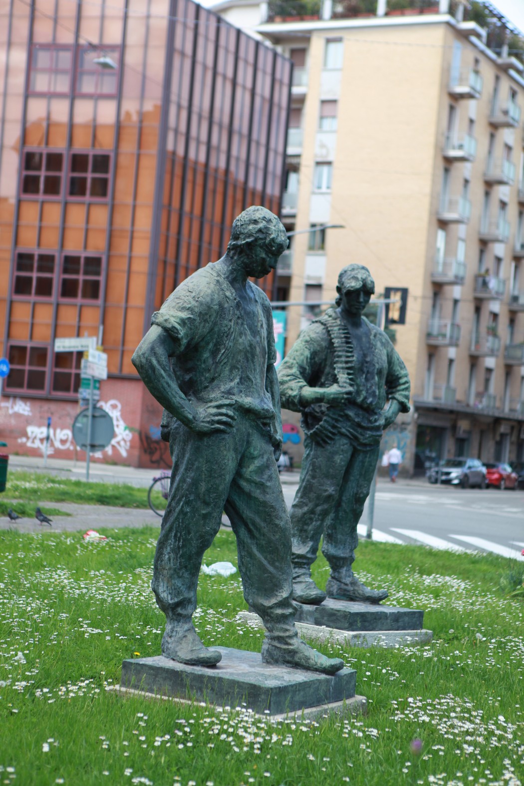 Due sculture bronzee di partigiani in piedi, con espressioni risolute, su un prato circondato da edifici urbani.