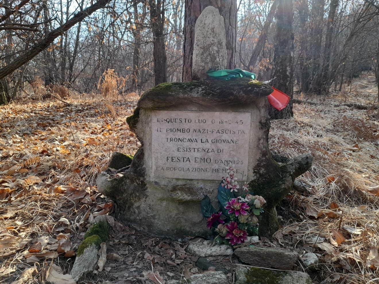 Cippo commemorativo in un bosco, dedicato alla giovane vittima del regime nazifascista, adornato con fiori.