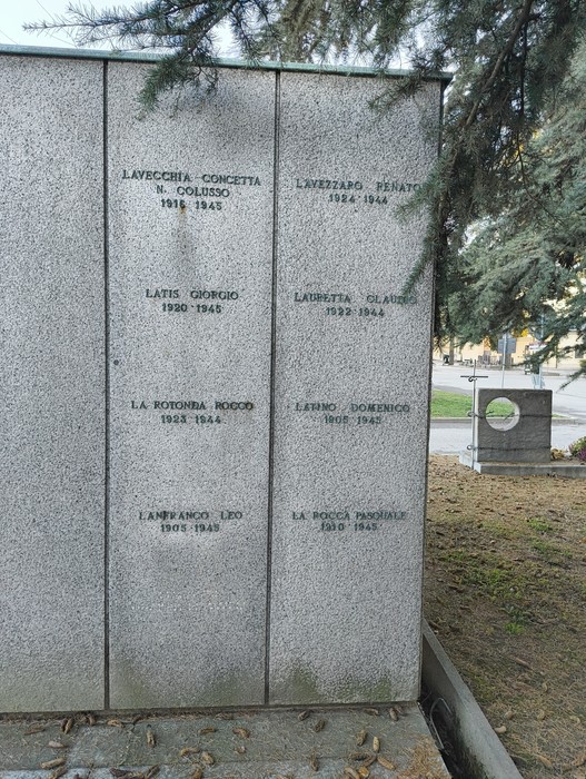 Monumento in granito con nomi di caduti, circondato da alberi e un'area verde. Atmosfera di commemorazione.