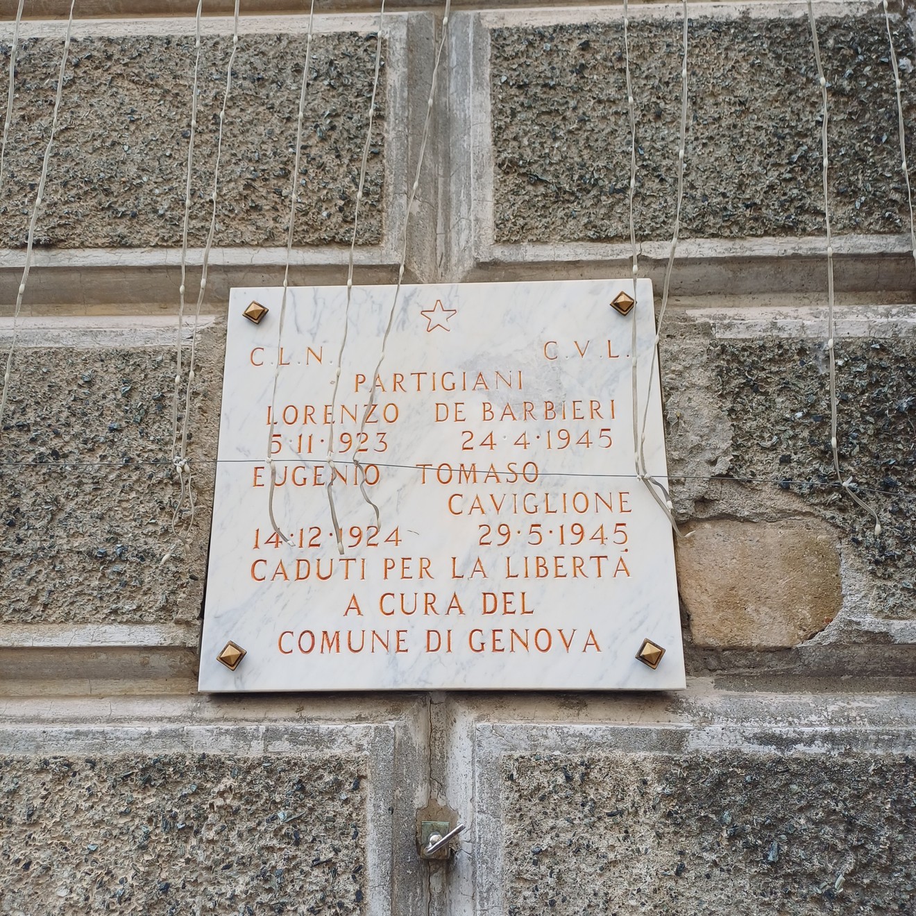 Lapide marmorea con iscrizione, fissata a parete in pietra con viti visibili agli angoli.