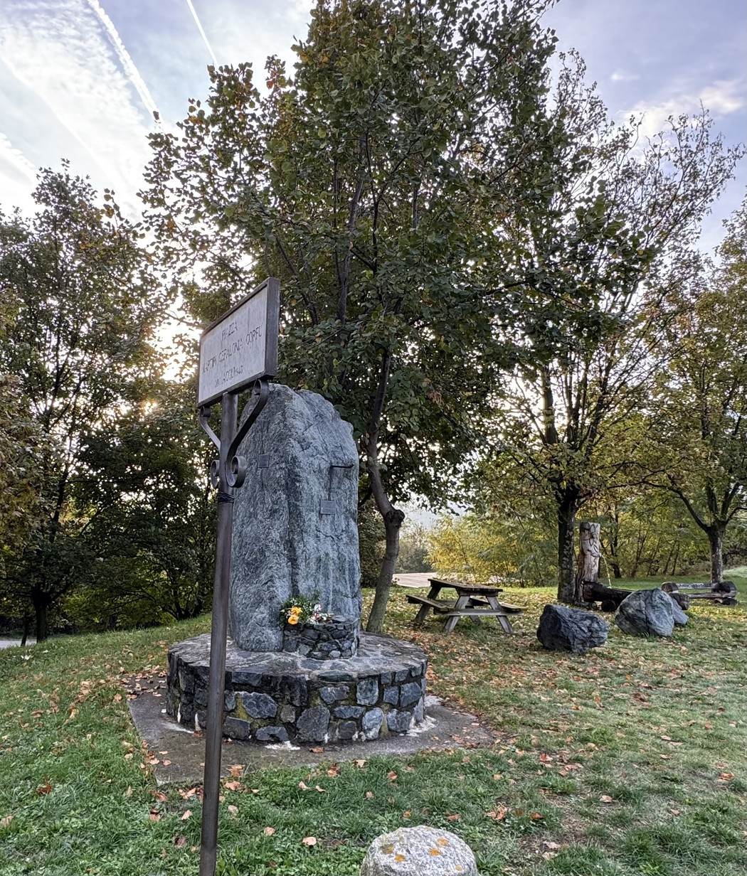 Stele in pietra su basamento circolare in un parco, con targa e iscrizione, circondata da alberi.