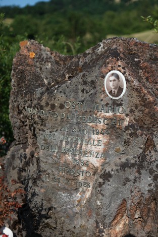 Monumento a Osti, commemorativo con foto e iscrizioni, situato in un contesto naturale.