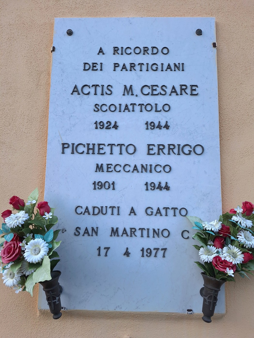 Lapide marmorea su parete, con iscrizione dedicatoria, decorata ai lati da due composizioni floreali artificiali.