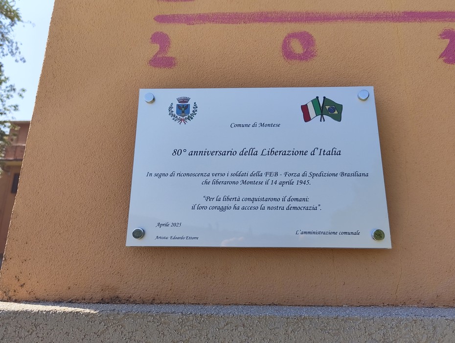 Targa bianca con stemma comunale e bandiere, fissata su una parete esterna, con iscrizione commemorativa.