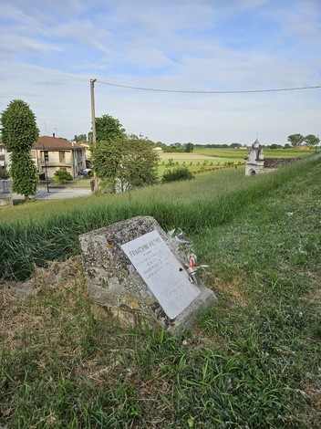 Cippo commemorativo situato in un paesaggio rurale con edifici e vegetazione circostante.