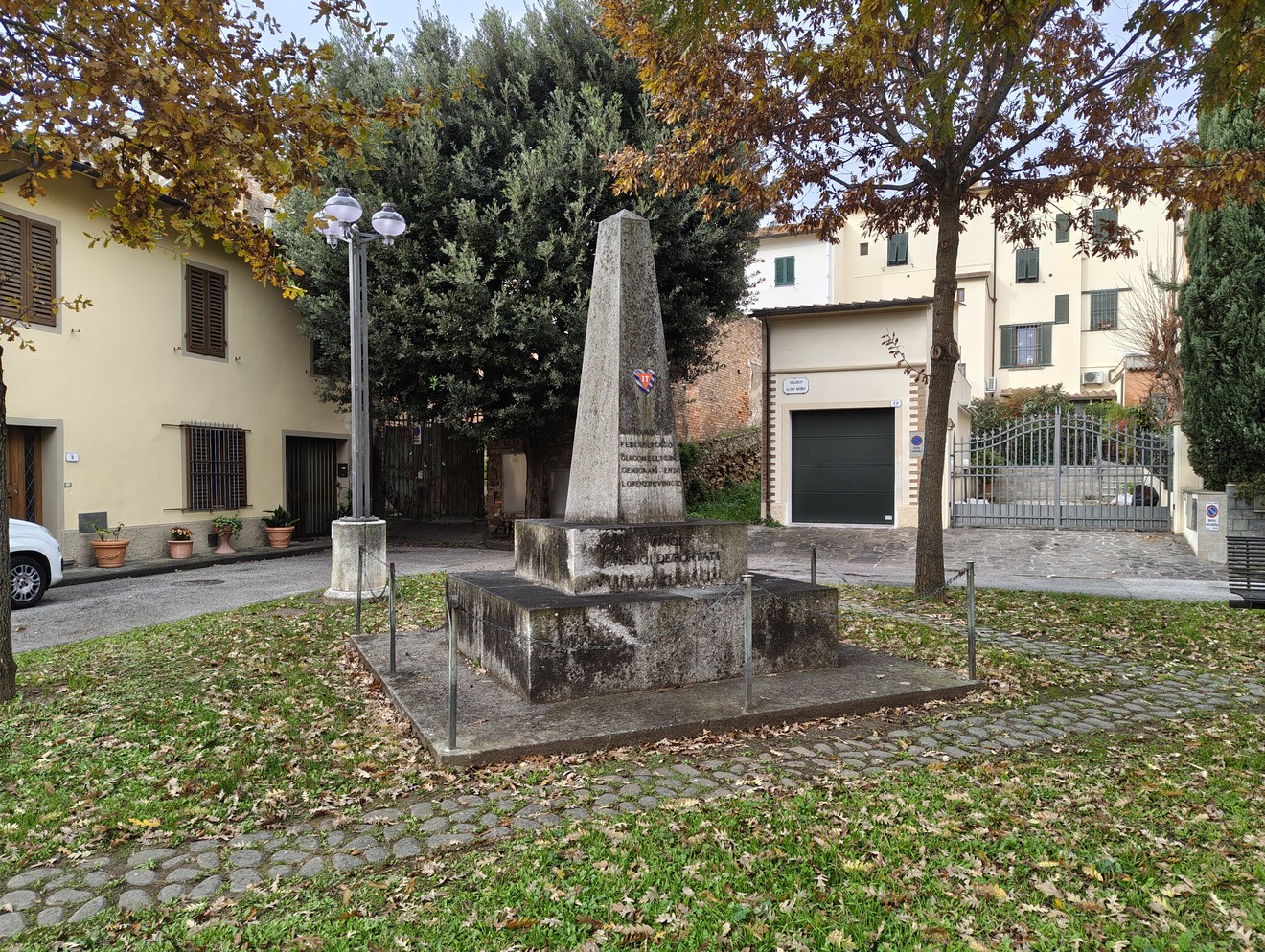 Monumento ai deportati situato in un'area verde, circondato da alberi e case, con una struttura obelisco.