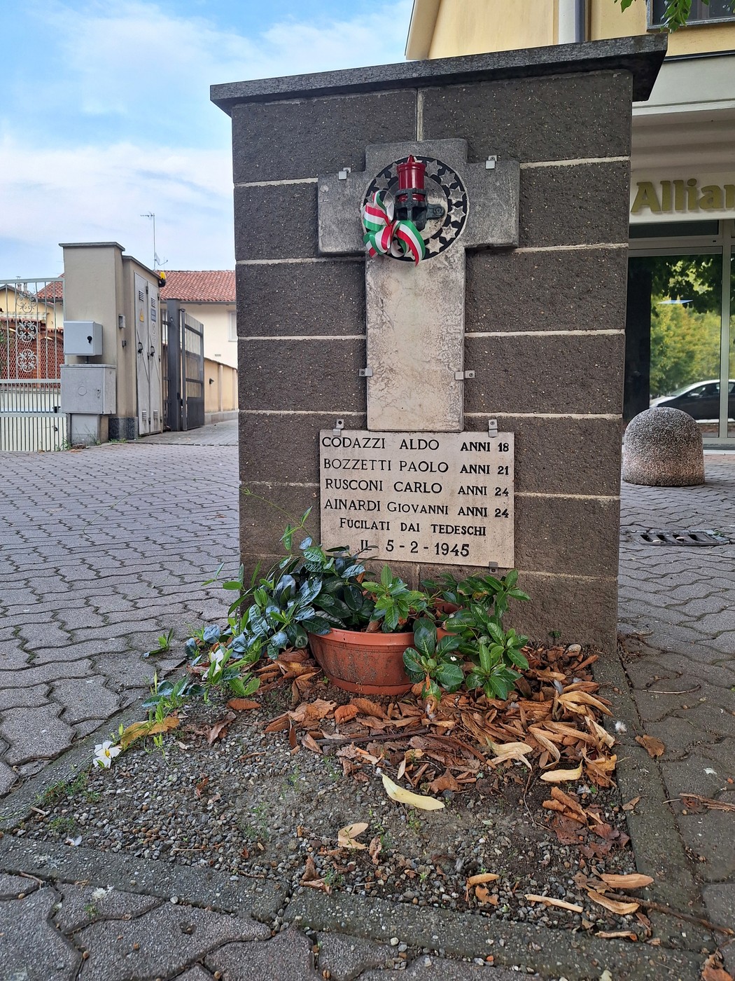 Cippo in pietra su marciapiede cittadino, con croce, stemma e targa con iscrizione, fiori ai piedi.