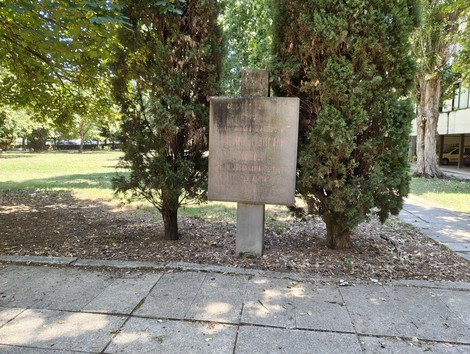 Stele in pietra con iscrizione, su basamento, tra alberi di cipresso in un'area verde.