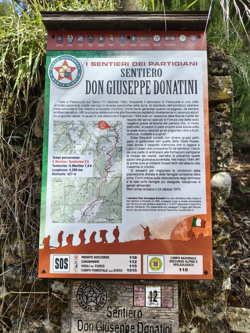 Pannello informativo illustrato nel bosco, con mappa, testo esplicativo e numeri di emergenza sul sentiero Donatini.