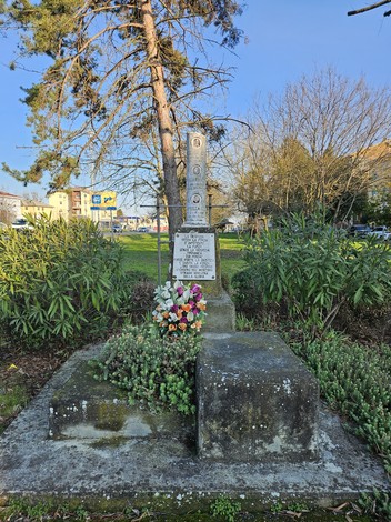 Cippo commemorativo circondato da vegetazione e fiori, con un albero sullo sfondo e un cielo sereno.