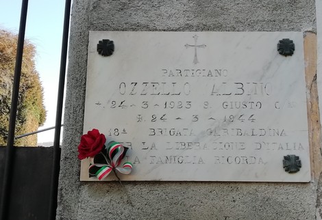 Lapide commemorativa di Albino Ozzello, partigiano, con decorazioni floreali e tricolore.