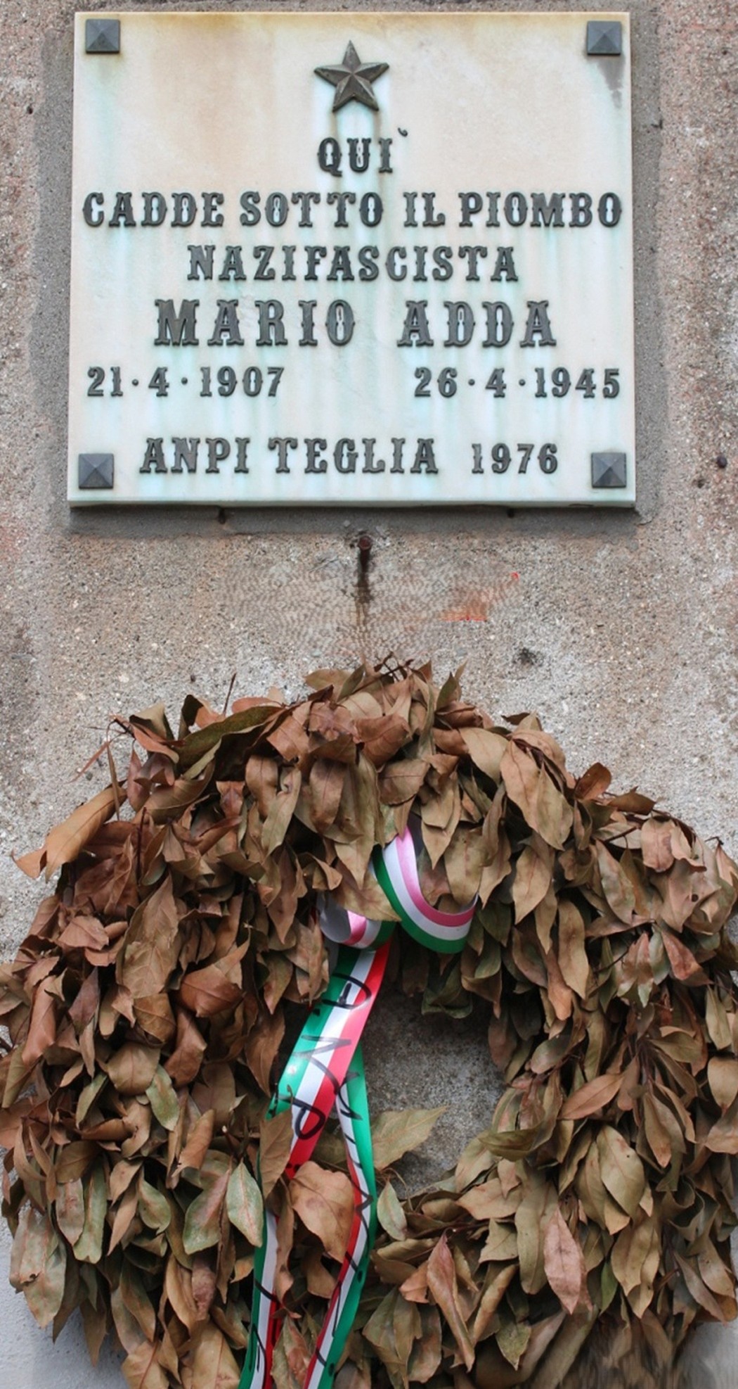 Lapide commemorativa di Mario Adda, nazifascista, con corona di alloro e nastri tricolori sullo sfondo.