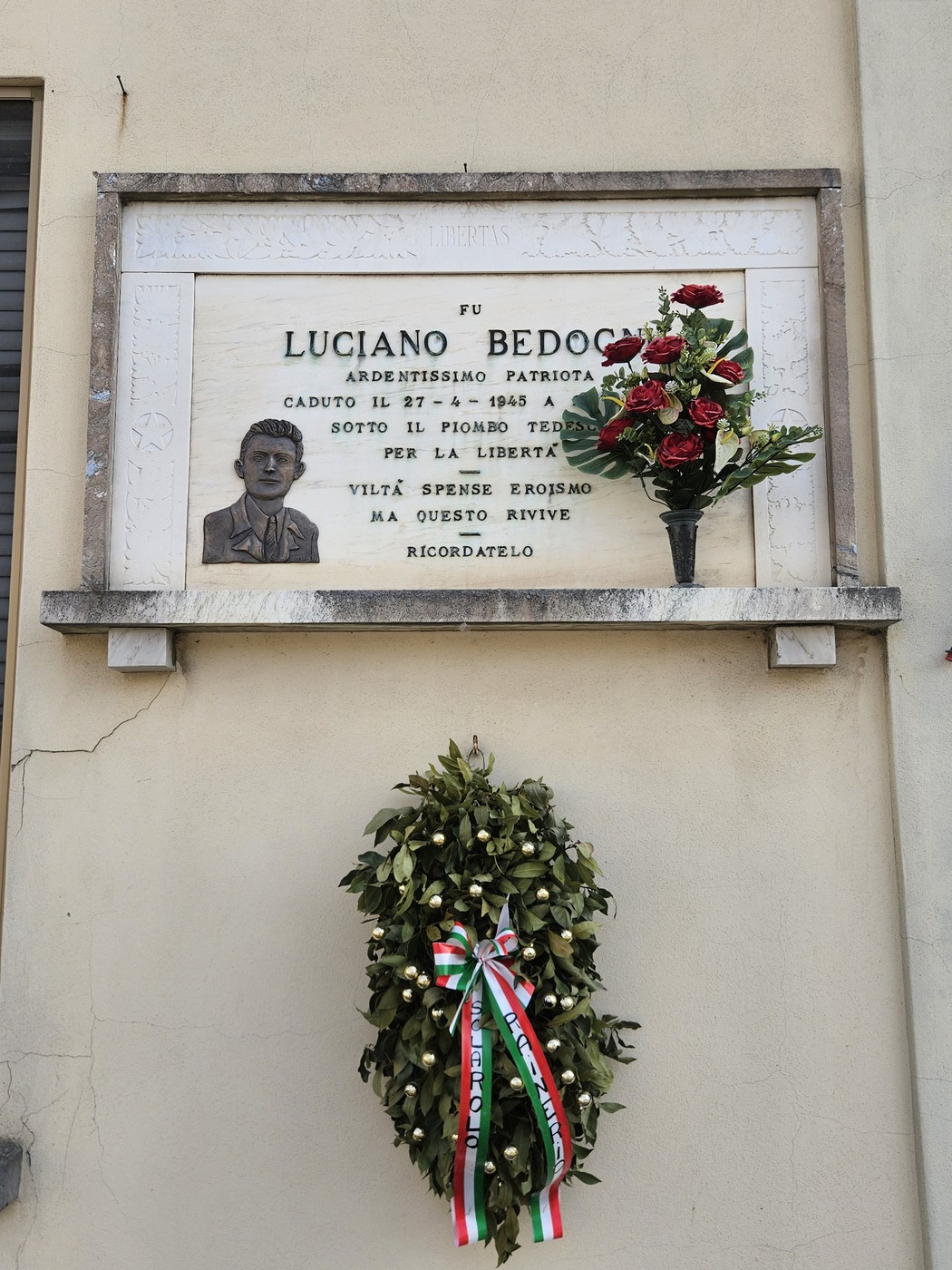 Lapide commemorativa di Luciano Bedogni, con fiori e corona, dedicata al patriota caduto per la libertà.