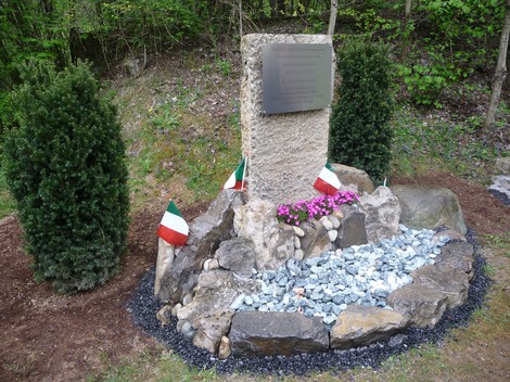 Cippo commemorativo ad Adobati, circondato da pietre, fiori e bandiere italiane, immerso nella natura.