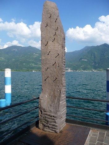 Monumento ai Tredici Martiri, stele in pietra con chiodi, affacciato su un lago e circondato da montagne.