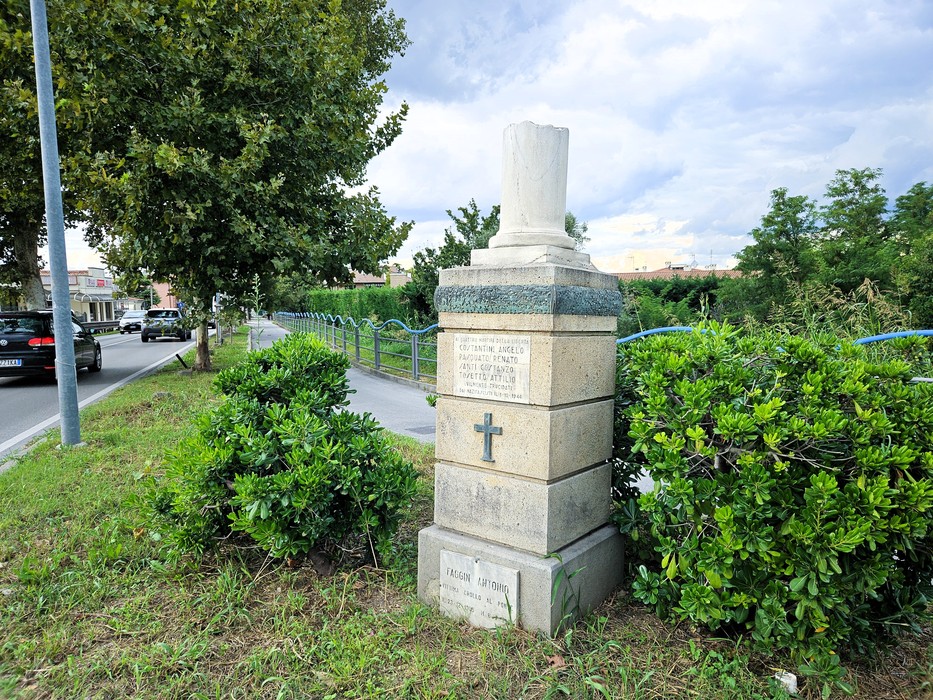 Cippo in pietra con base a gradoni e colonna spezzata, iscrizione incisa, ai margini di una strada alberata.