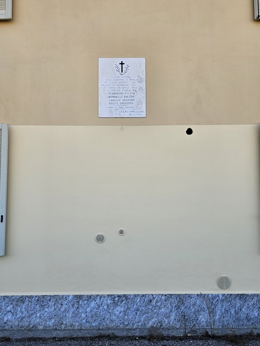 Lapide marmorea con croce incisa e iscrizione, affissa su parete esterna color ocra di un edificio.