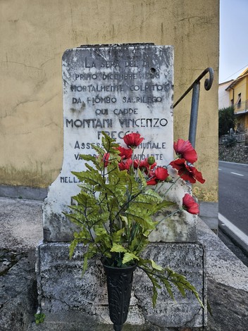 Cippo commemorativo dedicato a Montani Vincenzo, con fiori rossi, situato lungo la strada.