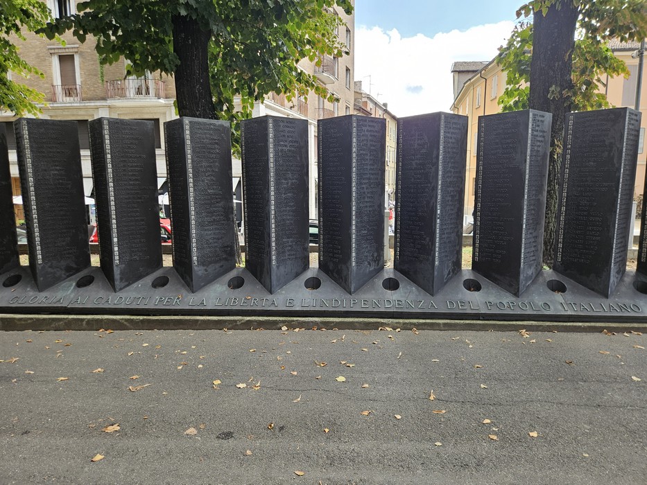 Monumento in metallo con pannelli incisi, disposto lungo una strada alberata.