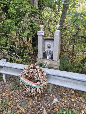Cippo in pietra con fotografia e iscrizione, guardrail e corona di foglie secche davanti, nel bosco.