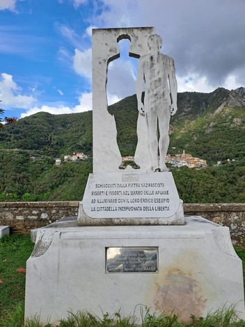 Monumento in marmo bianco con sagoma ritagliata e figura umana scolpita, su basamento con iscrizione, tra i monti.
