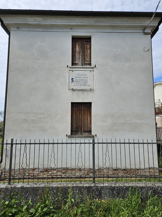 Lapide a Mazzo su un muro di una casa, con finestre chiuse e recinzione in metallo.