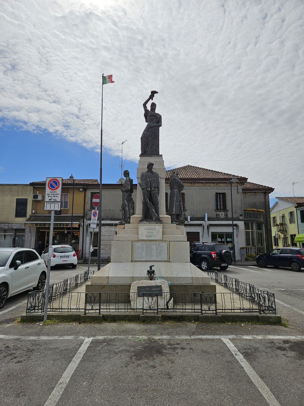 Monumento ai Caduti di Grignano Polesine, con statue in bronzo e base commemorativa, circondato da edifici e una bandiera.
