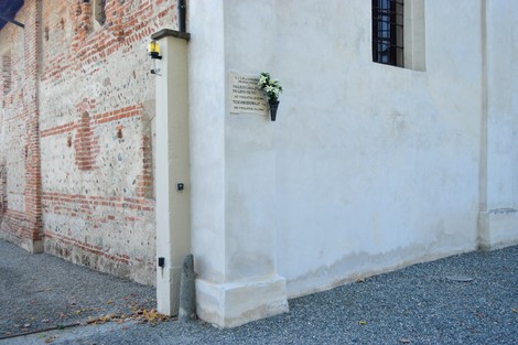 Lapide commemorativa nei pressi di un edificio storico, con fiori decorativi accanto.