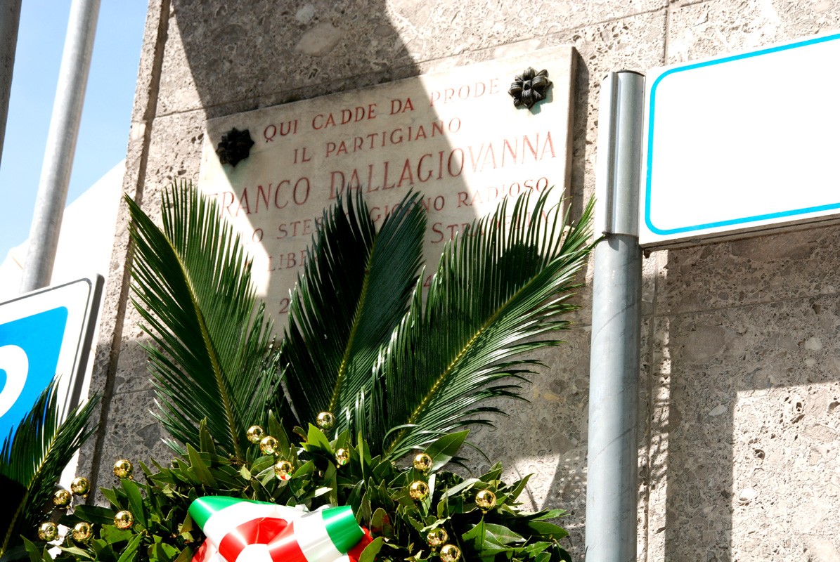 Lapide commemorativa di Franco Dellagiovanna, partigiano, con decorazioni floreali e segnaletica stradale.
