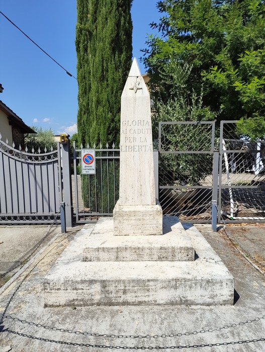 Cippo in pietra a forma di obelisco con iscrizione, su basamento a gradini tra cancelli in un giardino.
