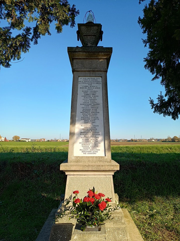 Monumento ai caduti dell'Amola, con iscrizioni e fiori rossi, immerso in un paesaggio sereno e verdeggiante.