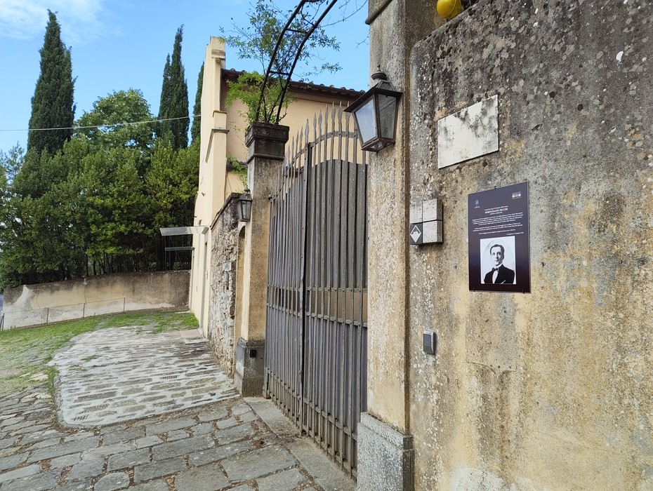 Ingresso con cancello in ferro battuto e pannello informativo su un muro, circondato da vegetazione.