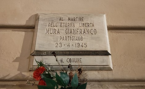 Lapide marmorea con iscrizione fissata su una parete, con fiori deposti sotto, in ricordo di un partigiano.