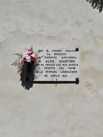 Lapide commemorativa dedicata a Aldo Quartieri, con fiori, su parete esterna.