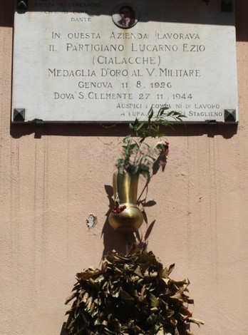 Lapide commemorativa dedicata al partigiano Lucarno Ezio con decorazioni floreali e foglie secche.