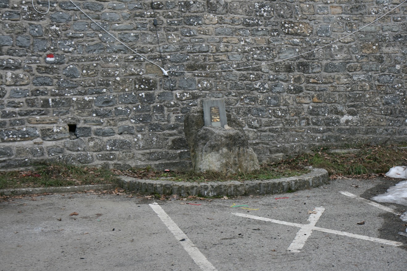 Cippo commemorativo di una rappresaglia, situato in un'area pubblica con una parete di pietra sullo sfondo.