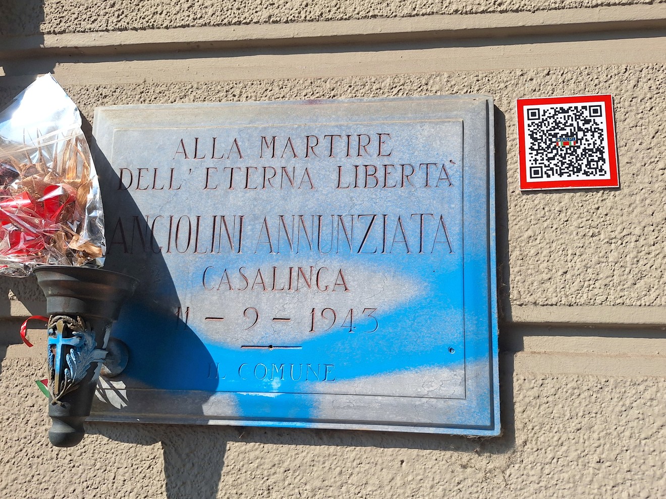 Lapide in marmo su parete esterna, con iscrizione e vernice blu versata sopra, accanto a un lampioncino con fiori.