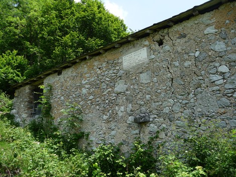 Lapide storica sulla parete di una cascina in pietra, circondata da vegetazione lussureggiante.