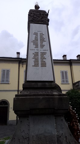Monumento ai Caduti con iscrizioni commemorative, situato in un contesto urbano, sotto un cielo nuvoloso.