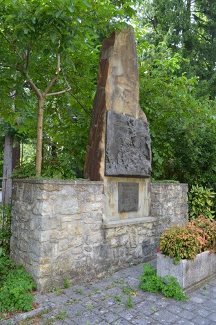 Monumento a Ricci, un obelisco in pietra circondato da vegetazione e una base decorata con un pannello.